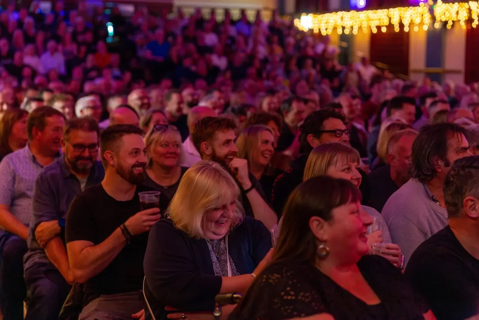 A seated audience laughing under purple lighting at a live comedy night.