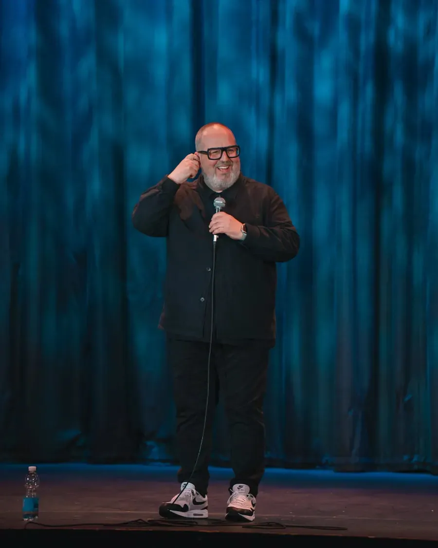 A comedian performing on stage in front of blue curtains.
