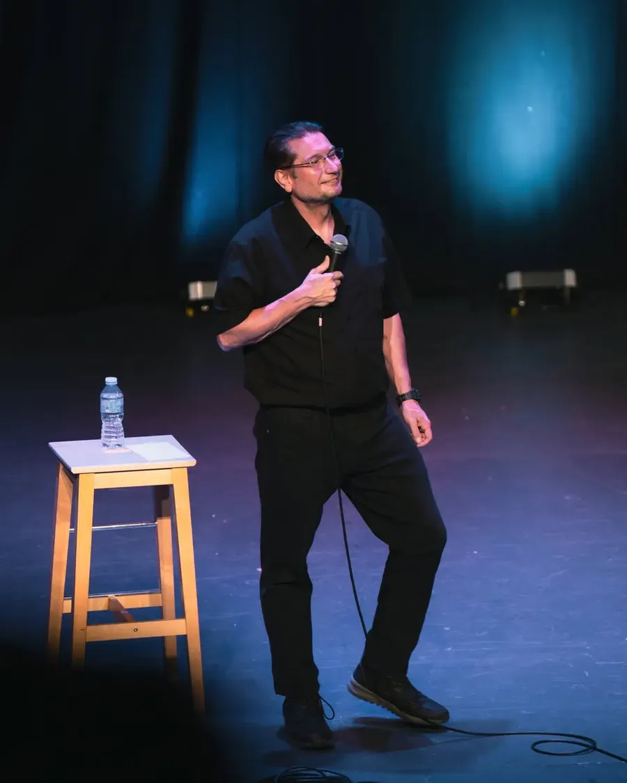 A comedian standing on stage beside a stool, holding a microphone.